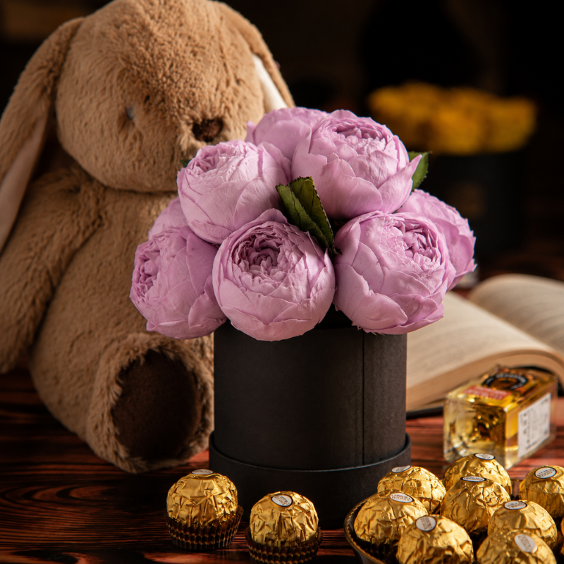 Arreglo de 14 peonías eternas en color morado lila montadas en sombrerera negra con chocolates de regalo