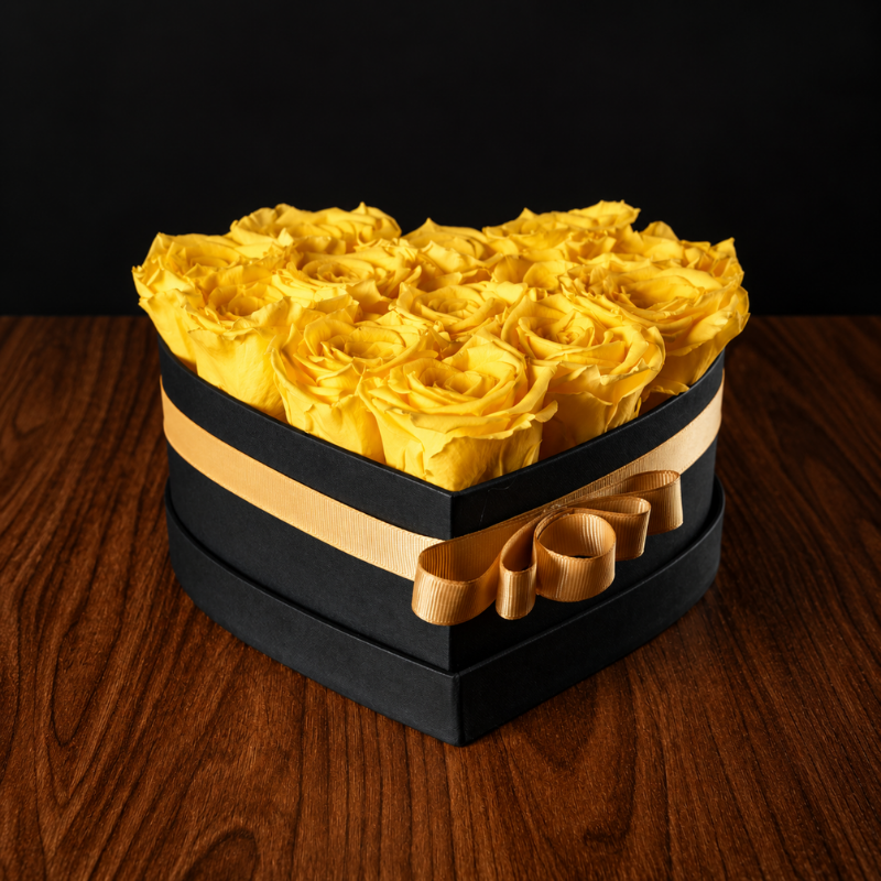 Caja de corazón negra con listón dorado y 13 rosas eternas amarillas preservadas sobre mesa de madera